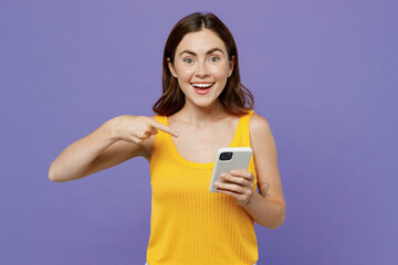 Young happy fun woman she wear yellow tank shirt hold use point index finger on mobile cell phone chatting in social network isolated on plain pastel light purple background. People lifestyle concept.