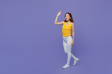 Full body side view young happy fun woman 20s she wear yellow tank shirt look camera walking go waving hand isolated on plain pastel light purple background studio portrait. People lifestyle concept.