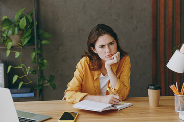 Young minded pensive thoughtful puzzled sad employee business woman 20s she wearing casual yellow shirt writing in notebook look camera sitting work at wooden office desk. Achievement career concept.