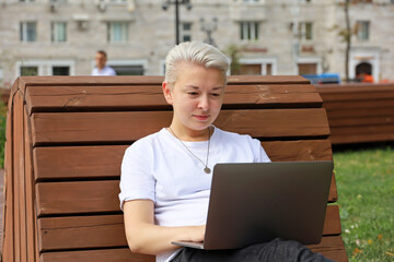 Girl with short blonde hair wearing white t shirt sitting on wooden bench with laptop on city...