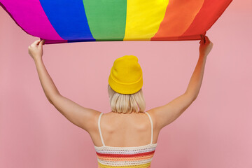 Back rear view young caucasian blond lesbian woman she wear yellow hat hold striped flag overhead isolated on plain pastel light pink background studio portrait. People lgbtq lifestyle fashion concept - Powered by Adobe