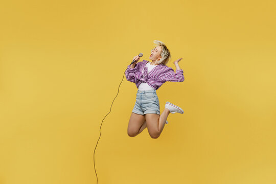 Full body young blonde woman 20s she wear pink tied shirt white t-shirt jump high sing song in microphone at karaoke club isolated on plain yellow background studio portrait. People lifestyle concept.