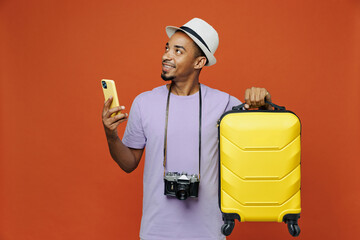 Traveler black man wear purple t-shirt hat hold suitcase mobile phone isolated on plain orange color background. Tourist travel abroad on weekends spare time getaway. Air flight trip journey concept.