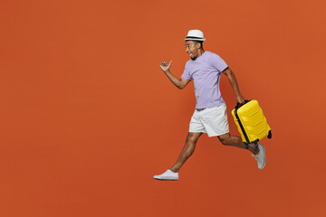 Full body traveler black man wear purple t-shirt hat jump high hold suitcase isolated on plain orange color background Tourist travel abroad in spare time rest getaway Air flight trip journey concept