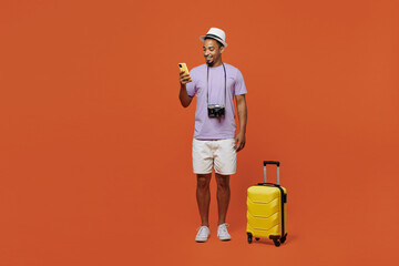 Full body traveler black man wear purple t-shirt hat stand with bag mobile phone isolated on plain orange background. Tourist travel abroad in spare time rest getaway. Air flight trip journey concept.