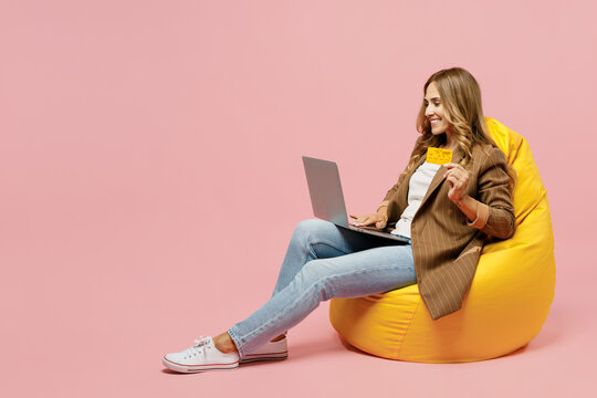 Full body young fun employee business woman she wear jacket sit in bag chair us laptop pc computer hold credit bank card shopping online order delivery isolated on plain pastel light pink background.