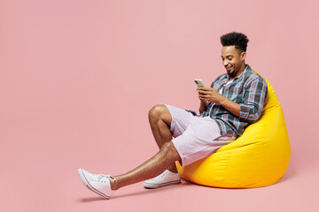 Full body young man of African American ethnicity 20s he wear blue shirt sit in bag chair hold in hand use mobile cell phone isolated on plain pastel light pink background. People lifestyle concept.