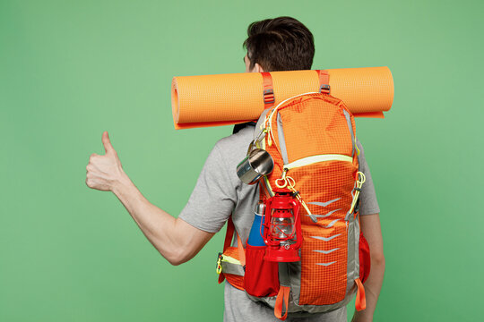 Back view young traveler white man carry backpack stuff mat show thumb up isolated on plain green background. Tourist leads active lifestyle walk on spare time. Hiking trek rest travel trip concept.