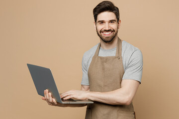 Young IT man barista barman employee wear brown apron work in coffee shop hold use laptop pc computer surfing internet isolated on plain pastel light beige background. Small business startup concept.