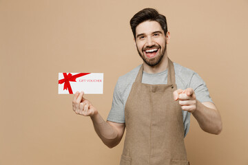 Young man barista barman employee in brown apron work in coffee shop hold gift coupon voucher card point finger camera on you isolated on plain light beige background Small business startup concept