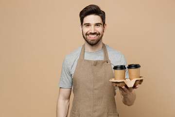 Young smiling happy man barista barman employee wear brown apron work in shop hold craft paper brown cup coffee to go isolated on plain pastel light beige background. Small business startup concept.