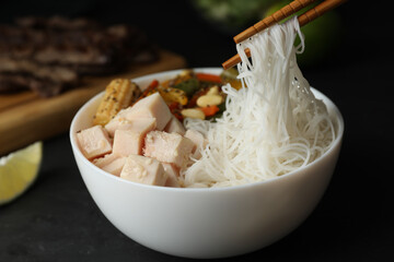Chopsticks with tasty cooked rice noodles over bowl on black table, closeup