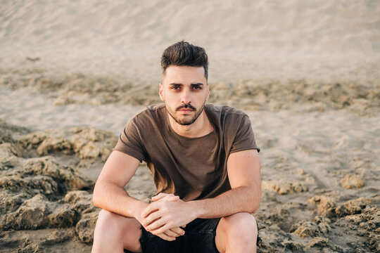 Handsome young Hispanic man sitting on sandy beach clasping hands