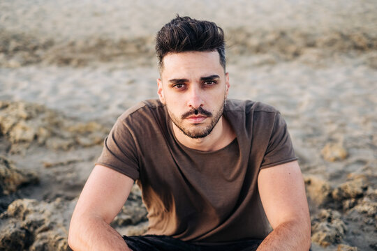 Handsome young Hispanic man sitting on sandy beach clasping hand