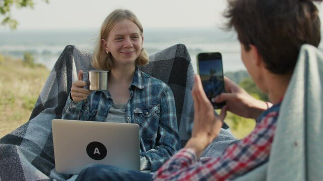 Man taking picture of woman sitting on camping chairs and working outdoors. Couple using laptop and mobile phone on picnic.