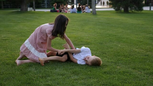 Funny girl tickles a boy in the park on the green grass in the evening at sunset