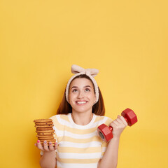 Portrait of smiling woman wearing striped shirt and hair band holding red dumbbell and cookies,...
