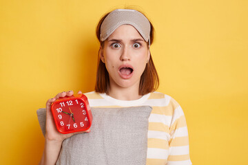 Shocked surprised woman wearing striped shirt and sleeping mask, holding pillow and red alarm...