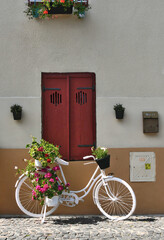 Fototapeta na wymiar white bicycle, flowered of geraniums, under a window whose red shutters out of wood are closed. small pots of flowers, a mailbox and a small door of iron dress the unit.