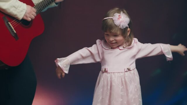 Little girl on stage in pink vintage dress, she dances, her father plays acoustic guitar. Color music is shining. Performance on stage. Early development of children
