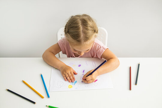 Blonde child girl draws with colored pencils sitting at the table. Top view, flat lay