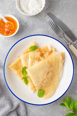 Stack of traditional thin pancakes or crepes on a plate with honey and sour cream on a gray concrete background. Food for Maslenitsa. Copy space