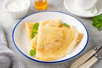 Stack of traditional thin pancakes or crepes on a plate with honey and sour cream on a gray concrete background. Food for Maslenitsa. Copy space