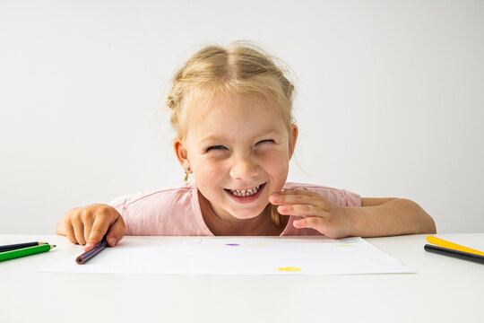 A smiling child blonde girl draws with colored pencils sitting at a white table