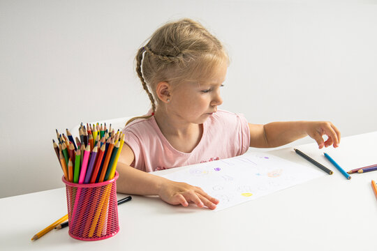 A blonde child girl draws with colored pencils sitting at a white table