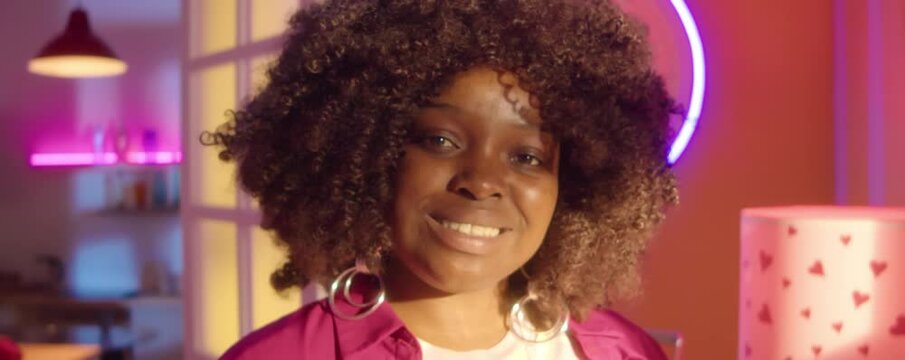 Portrait of African American party girl wearing fuchsia outfit and y2k jewelry smiling on camera while posing in room with disco lights and neon decorations