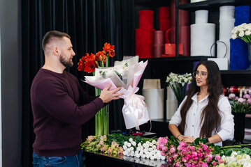 A young bearded man buys a beautiful bouquet of flowers for a girl's holiday in a cozy flower shop. Floristry and bouquet making in a flower shop. Small business.