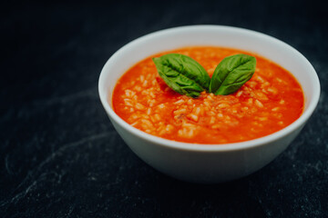 Tomato soup with rice and basil in a white bowl on a dark stone table