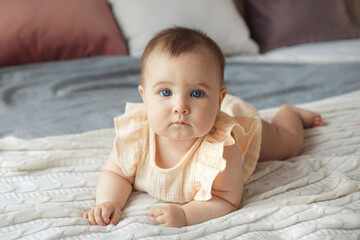 Cute little baby girl in cotton dress with blue eyes lying on bed and looking at camera