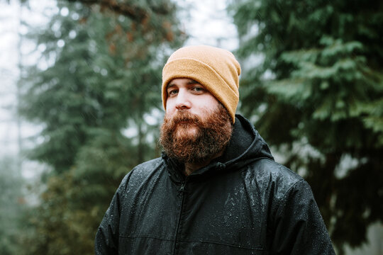 Bearded pensive guy in coat and hat near fir trees
