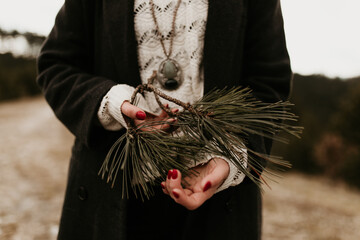 A woman is holding a pinewood branch with leaves in her hands. She is wearing white lace sweater, a black coat and a beautiful necklace in the background. She is gently holding the plant, copy space.