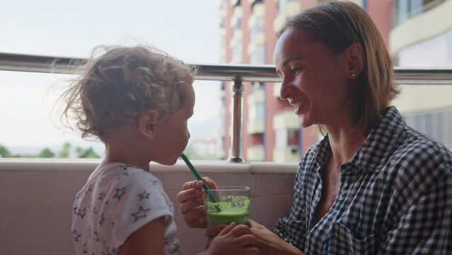 Caucasian baby sitting on the kitchen table among green leaves drinking green smoothie they've just prepared with its young mother. High quality 4k footage