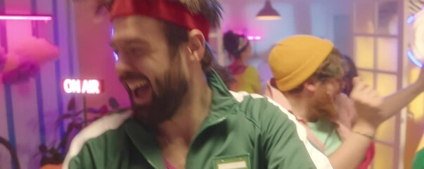 Young bearded man wearing green tracksuit and fitness headband performing energetic dance at 90s throwback party with friends in room colorful disco lights