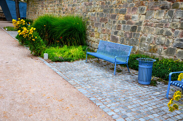 blue metal park bench under a brick wall in the park. soft sandy park paths and beds of tall yellow...