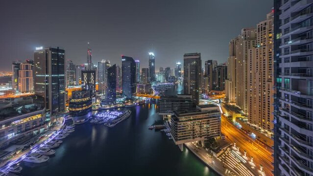 Aerial view to Dubai marina illuminated skyscrapers around canal with floating yachts during all night timelapse with lights turning off. White boats are parked in yacht club