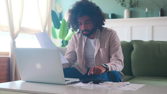 Young focused ethnic Arabian man works with financial documentation and calculator auditing tax that need to be paid to government agencies sits on sofa in living room. Business, economics, taxation