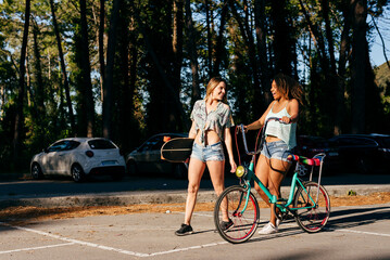 Female friends walking on street holding skate board and bicycle