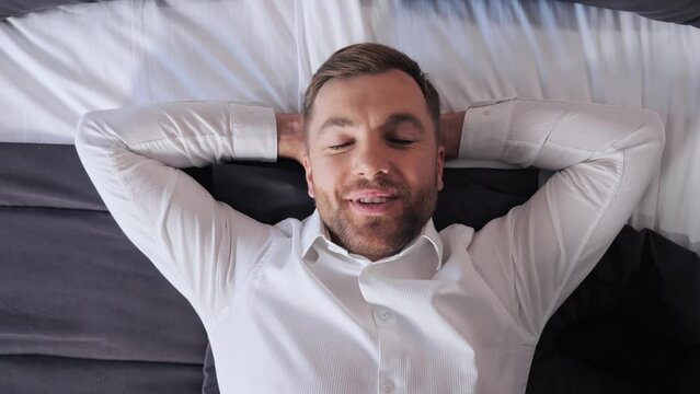 Man lying on the bed relaxing after a hard day, enjoying a soft mattress and pleasant linen textiles. Businessman rest at daytime. Handsome man in white shirt feeling good in comfortable hotel. 