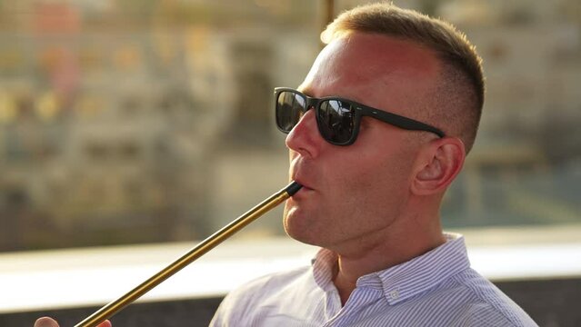 Blond man wearing sunglasses smoking hookah. Man produces smoke from nose and mouth at once. Close up.