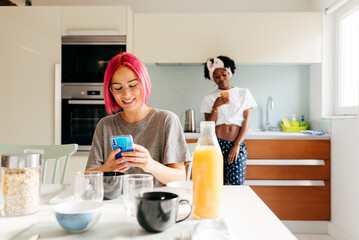 Cheerful young woman using smartphone near cooking black friend
