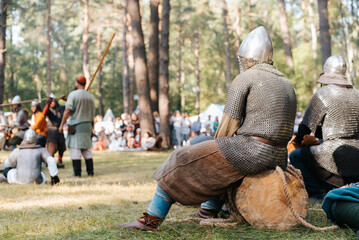 Festival of historical reconstruction and medieval costume performances in open air. Back view of warrior in helmet and chain mail resting on log and watching battle of spearmen on battlefield - obrazy, fototapety, plakaty