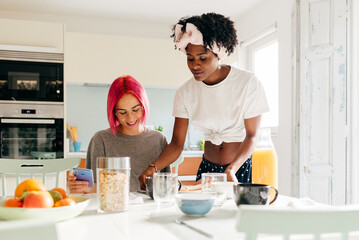 Cheerful young woman using smartphone near cooking black friend