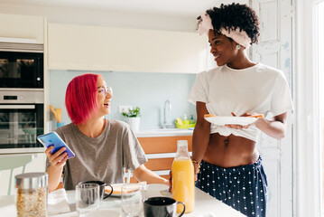 Cheerful young woman using smartphone near cooking black friend