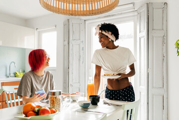 Cheerful young woman using smartphone near cooking black friend