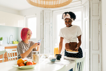 Cheerful young woman using smartphone near cooking black friend