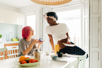 Cheerful young woman using smartphone near cooking black friend